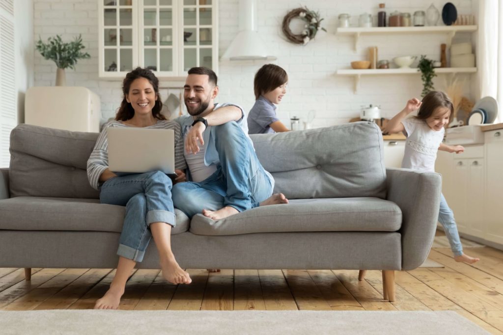 Junge Eltern sitzen mit dem Laptop auf dem Sofa. Die Kinder rennen um das Sofa herum